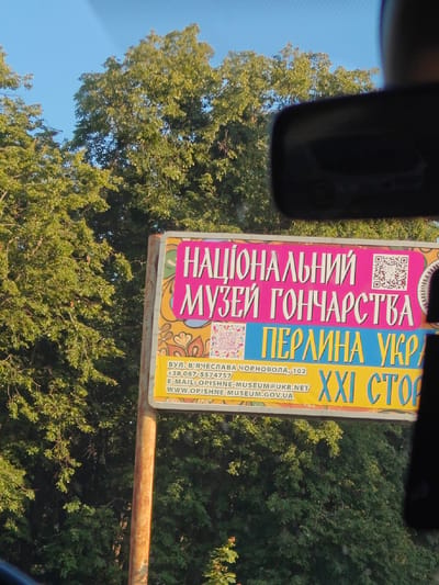 The image shows a billboard with Ukrainian text and a QR code. The text on the billboard appears to be advertising the National Museum of Pottery in Opishnia, Ukraine. The billboard features bright colors, with the main text in pink and yellow. Below the main text, there is contact information including a street address, phone number, email, and website. The background of the image is dominated by lush green trees and a clear blue sky. The billboard is mounted on a metal pole. The perspective suggests the photo was taken from inside a vehicle, possibly looking through a windshield, as a rearview mirror is partially visible in the upper right corner.