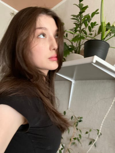 The image is a close-up, profile view of a young woman with long, dark brown hair. She is wearing a black t-shirt. Her head is turned to her left, and she is looking upwards and to her left. Her facial features are visible, including her nose, lips, and eyes, with a few freckles on her nose and cheek. Her skin appears fair. In the background, to her right, there is a white shelf holding two potted plants. One plant is dark green and leafy in a black pot, and the other is a lighter green vine with small leaves in a white pot. The wall behind the shelf is a neutral, textured color. A thin white rope or cord hangs vertically in the right side of the frame. The lighting appears to be soft and natural. There are no indications of the specific location in Nizhnyaya Maktama, Russia.
