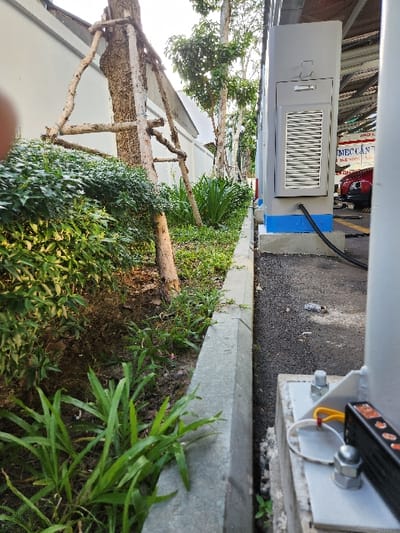 A grey metal utility box with a ventilation grille is positioned next to a curb. A black cable extends from the box. Adjacent to the box is a grey electrical component attached with bolts. Below, lush greenery and vegetation fill the space between the structure and a white wall. A rustic wooden structure supports the plants. The scene suggests an urban or suburban outdoor setting.