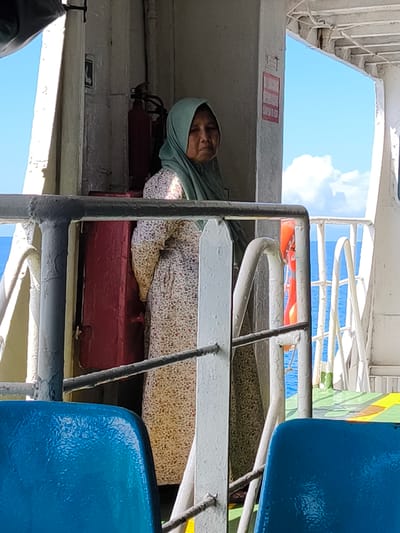 On a ferry in Gunung Malang, Indonesia, a middle-aged woman wearing a light green hijab and a floral print dress stands behind a railing. Behind her, a red fire extinguisher is mounted on a wall. Through the ferry's open side, the blue ocean and sky are visible with some white clouds. In the foreground, the tops of two blue plastic chairs are partially visible.