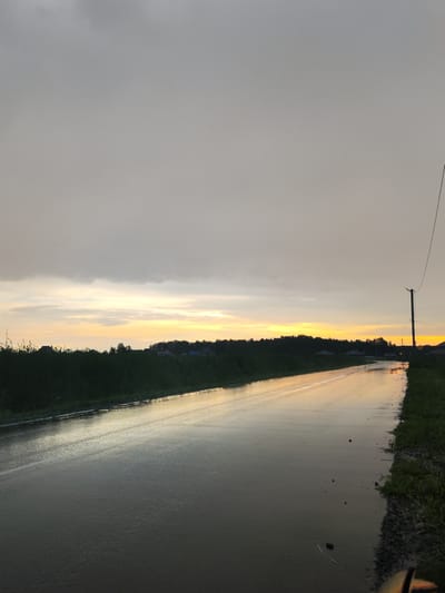 This media file captures a scene at dusk, likely in the vicinity of Paderina, Russia. The image depicts a wet, rural road reflecting the warm hues of a sunset, suggesting recent rainfall or damp conditions. The sky is overcast, with a band of orange and yellow light breaking through near the horizon, indicating the sun is setting.
The road itself is the central focus, stretching into the distance and appearing slick with water. To the left of the road, a dense line of dark green foliage, possibly trees and bushes, creates a natural border. Beyond this greenery, hints of scattered houses and structures are visible, suggesting a small settlement or rural community. To the right of the road, the terrain is covered in grass and gravel, with a solitary utility pole standing tall, its wire extending across the frame. The overall mood is one of quiet solitude and the serene beauty of a fading day. No people or specific objects are prominently featured, allowing the natural elements of the landscape to dominate the composition. The time of day is clearly evening, transitioning into night, with the weather appearing to be post-rain.
