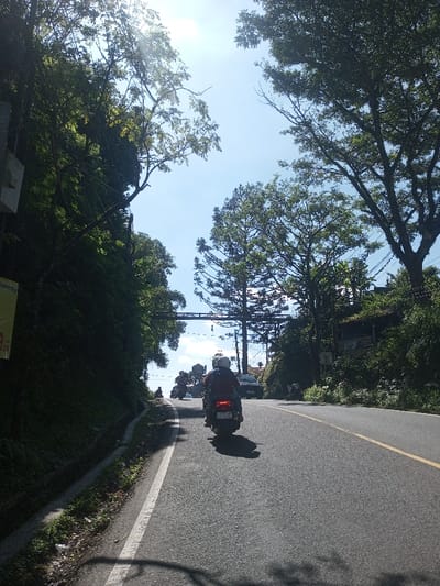 The scene captures a sunny day on a paved road in Ban Phonhxavanh, Laos. A dark-colored scooter, ridden by an individual wearing a white helmet and dark jacket, is central, traveling uphill away from the viewer. Its red taillight is active, and a license plate is partially discernible. Ahead, a line of at least four additional scooters and a dark car proceed in the same direction. The asphalt road, marked by a solid white line on the left shoulder and a double yellow line further ahead, curves gently to the right. To the left, a dense, verdant embankment rises steeply, partially obscuring a light-colored sign and a yellow banner. The right side of the road is bordered by abundant trees, utility poles, and distant structures with brown roofs. A dark metal overhead gantry spans the road in the mid-ground. The bright blue sky shows significant sun flare in the upper left, casting distinct shadows onto the foreground road.