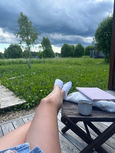 The image depicts a person's legs, clad in white socks and denim shorts, resting on a wooden deck. In the foreground, a small wooden table holds a mug of tea, a white knitted blanket, and a pink tablet. Beyond the deck, a vast expanse of green grass and trees stretches towards a dramatic sky filled with dark, ominous clouds. The setting appears to be rural, likely outside of Znamenka, Russia, given the presence of birch trees and dense foliage characteristic of the region. The overall atmosphere is one of quiet contemplation, juxtaposed with the impending stormy weather.