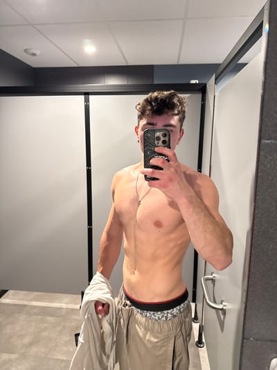 A male individual is depicted taking a mirror selfie within a locker room or changing facility in Chantepie, France. The subject, possessing a muscular physique, has short, dark, curly hair and is shirtless. He wears light-colored (khaki/tan) cargo-style pants, with a visible patterned underwear waistband featuring black and red trim. A thin silver chain with a small pendant is worn around his neck, and a dark stud earring is visible in his left ear. He holds a black smartphone with a white topographic-patterned case in his right hand, which obscures the lower portion of his face. His left hand holds a crumpled white garment, possibly a t-shirt or towel. The background comprises light gray partition panels, indicative of changing stalls, separated by a dark vertical beam. To the right, a darker gray wall is visible, equipped with a silver horizontal grab bar. The floor is tiled in a light gray, and the white ceiling contains recessed lighting fixtures.