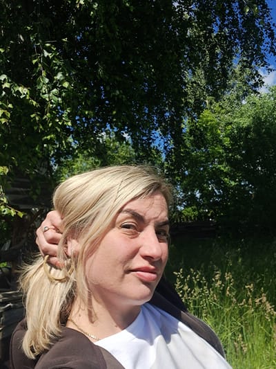 A bright, sunlit, outdoor portrait features a fair-skinned woman with blonde hair, visible from her upper chest to the top of her head. She is looking directly at the camera with a neutral to slightly smiling expression. Her blonde hair is styled loosely, with some strands falling around her face, and one hand is raised, possibly touching her hair or face. She wears a white t-shirt under a dark, possibly brown or black, zippered jacket. A delicate gold chain necklace is visible around her neck, and a ring is on her finger.
The background is dominated by dense green foliage of trees, suggesting a natural setting like a park or forest. Patches of clear blue sky with some white clouds are visible through the gaps in the leaves. To the right side of the image, behind the woman, there's an area of tall, light brown and green grass or reeds, indicating a wilder or less manicured landscape. The strong sunlight illuminates the woman's face and hair, creating highlights and casting some gentle shadows, suggesting it is daytime, likely morning or late afternoon. No text is visible in the image. The scene is set in an outdoor, rural or natural area.