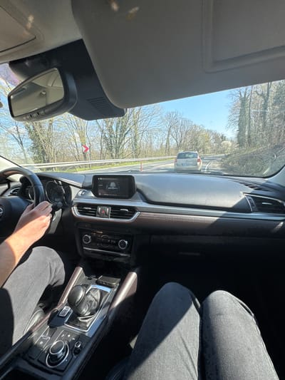 The image depicts the interior of a car, a Mazda CX-5, driving on a road with trees lining the sides, likely in Staro Oriahovo, Bulgaria, based on the license plate of the car in front (presumably Bulgarian). The dashboard is visible, displaying information on the car's infotainment screen. The driver's hand is visible on the steering wheel, and the driver is wearing dark colored pants. The sun is shining. The interior of the car is partly leather.