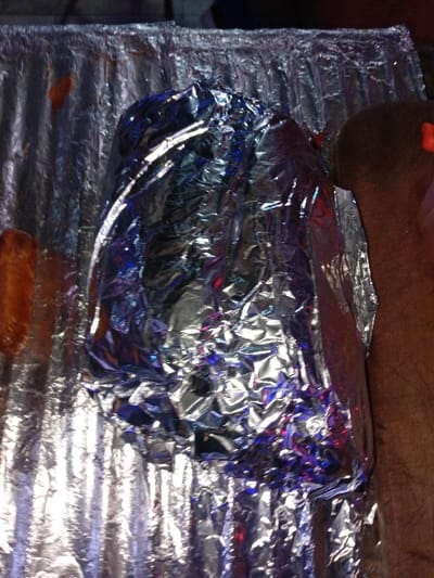 A package wrapped in aluminum foil sits on more foil. The foil has many wrinkles and reflects light, creating a shiny and uneven surface. Part of a tan-colored object is visible above the foil-wrapped package, likely resting on it. A small, brown, cylindrical object lies at the bottom of the frame, seemingly separate from the main package.