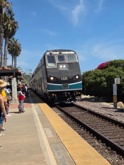 In San Clemente, California, a double-decker commuter train, numbered 643, is stopped at a station under a clear blue sky. The train, predominantly black with a teal stripe, faces the camera. A group of people, dressed in casual summer attire, are waiting on the platform. One person in the foreground wears a straw hat and yellow shirt, while others are carrying bags. Palm trees and lush green bushes with vibrant pink flowers are visible in the background, with a hillside dotted with buildings rising behind them. The train platform features a yellow textured safety strip along the edge. A sign on a post reads "NO DUMPING TRESPASSING." The scene captures a moment of transit at a sunny, coastal train station.