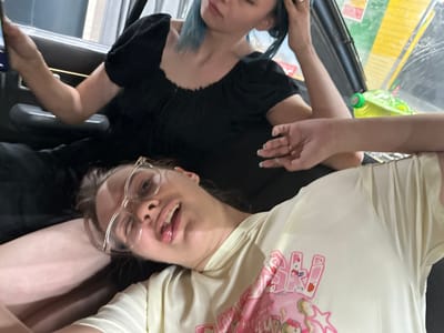 This image shows two people in what appears to be a casual setting, possibly a vehicle. One person is wearing a light-colored t-shirt with a graphic print featuring pink characters and text. This individual is looking towards the camera with their tongue sticking out slightly and wearing clear-rimmed glasses. The other person, mostly obscured, is wearing a black t-shirt. Their hair has streaks of blue.

A bright green plastic bottle with a green cap is visible in the foreground, resting on a surface that appears to be a car's interior. The background is dark and out of focus. The lighting suggests it is daytime, and no specific weather conditions are discernible. The overall impression is one of a candid, informal moment.

The text on the t-shirt appears to include some stylized letters, possibly "M" and "NG," along with some smaller, less legible words that could be English. The graphic on the shirt also includes star shapes and cartoonish characters, which might be related to anime or a similar style. The scene is likely indoors or in a shaded area, given the lighting and reflections.