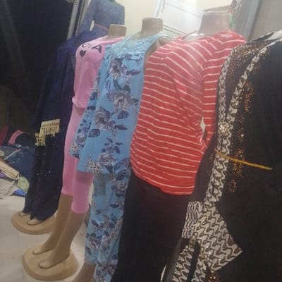 The image displays an indoor retail scene in Dan Maliki, Nigeria, featuring five headless mannequins arranged diagonally from left to right. The first mannequin, partially visible on the far left, wears a dark blue garment with gold fringed trim. The second mannequin wears a pink long-sleeved top featuring white lettering, possibly "CIA," paired with pink capri leggings. The third mannequin showcases a light blue two-piece ensemble with a floral pattern and a lace-trimmed neckline. The fourth mannequin displays a red and white horizontally striped long-sleeved top combined with black leggings. The fifth mannequin, partially visible on the far right, wears a black long dress or abaya adorned with intricate white and gold patterned embellishments and a thin yellow string tied at the waist. All visible mannequins are light brown in coloration, include hands and feet, and rest on rounded, light brown display bases. The background is out of focus, revealing blurred textiles and a light-colored wall with a white sign.