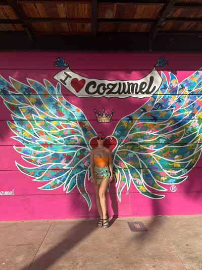 An adult female subject stands centered against a vibrant pink mural on an exterior wall in San Miguel de Cozumel, Mexico. The mural features a large pair of stylized wings with white outlines, filled with numerous small, colorful silhouettes of animals. The left wing contains marine life such as turtles and fish, while the right wing shows birds and various terrestrial creatures. Above the wings, a white banner held by two blue birds reads "I ❤️ cozumel". A multicolored crown is depicted below the banner, between the wings. The subject wears an orange strapless crop top, a patterned green, yellow, and blue wrap skirt, black open-toed sandals, a red visor, and sunglasses. She stands facing the camera with her right leg straight and her left leg slightly bent. Strong overhead sunlight casts distinct shadows of the subject and an unseen structure onto the reddish-brown concrete pavement. A dark structural beam and wooden ceiling are visible at the top of the frame.