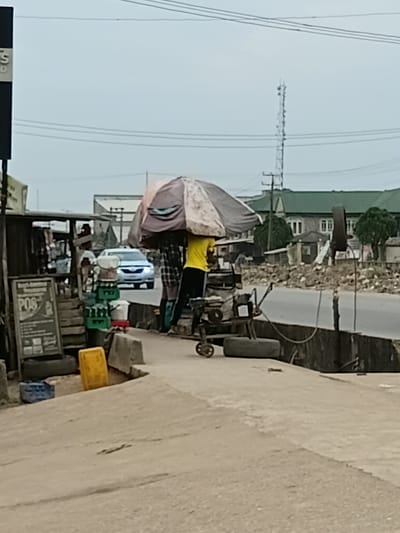 The image captures a street scene in Akute, Nigeria. A man wearing a yellow shirt stands under an umbrella. There is a motor vehicle on the road, utility poles, and buildings in the background. There is a kiosk on the side of the road.