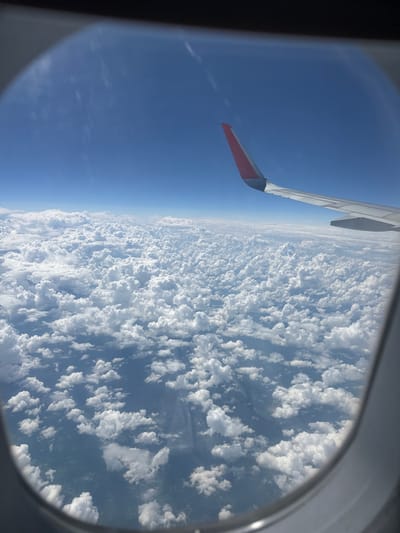 The image is a view from an airplane window, looking down at a vast expanse of clouds and the sky. A portion of the airplane's wing, with a red tip, is visible in the upper left quadrant, angled towards the right. The clouds below are fluffy and white, resembling a thick blanket. The sky is a clear, deep blue. The image appears to have been taken during the daytime, likely mid-morning or afternoon, given the bright lighting on the clouds and the clarity of the sky. There are no visible people or text in the image. The scene depicts a common experience of air travel, offering a perspective of the world from above.