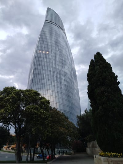 The image shows the Flame Towers in Baku, Azerbaijan, on a cloudy day. The skyscrapers are tall and modern, with a distinctive flame-like shape. They are covered in glass and reflect the sky. In the foreground, there are trees and a street with cars. There are also some people walking on the sidewalk. The overall impression is of a modern and bustling city.