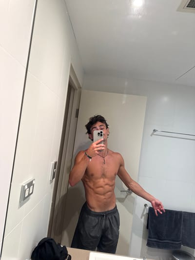 A male individual is depicted taking a mirror selfie in a bathroom located in Bondi, Australia. The subject, appearing in his late teens or early twenties, has dark, disheveled hair and light facial stubble. He is bare-chested, displaying a muscular physique with visible abdominal definition, and wears dark grey athletic shorts. On his left wrist are multiple bracelets, and around his neck, a dark necklace with a small pendant. He holds a light-colored smartphone with a three-lens camera system, which partially obscures his face, as he captures the image.

The bathroom environment features light-colored tiled walls and a white ceiling with a visible recessed light fixture and an air vent. To the left of the subject, two double power outlets with white faceplates and silver switches are mounted on the tiled wall. A light-colored door is visible slightly ajar in the reflection behind him. To the right of the subject, a silver metal towel rail holds two folded dark grey towels with lighter stripes. The bottom edge of a white basin or countertop is visible in the foreground, with a dark piece of fabric or clothing resting on it in the lower left corner. The scene is brightly illuminated by overhead lighting.