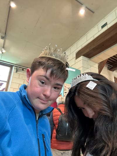 The image depicts two young adults, a male and a female, standing indoors at the Château de Chambord, a royal residence located in the Loire Valley, France, at coordinates 47.616 latitude and 1.518 longitude. The male, wearing a blue fleece jacket, is facing the camera, while the female, with long dark hair, is partially obscured by her hair, looking down. Both are wearing crowns on their heads; the male's crown is gold and the female's is silver. The male has a slight smile on his face. The image was captured in a white-walled room with a wooden beam overhead, and a white tile floor. In the background, a person, possibly a man, wearing an orange jacket and a backpack, is partially visible.