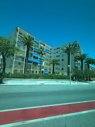 In La Pineda, Spain, a sun-drenched street scene unfolds with a clear blue sky overhead. In the mid-ground, two multi-story apartment buildings stand side-by-side. The building on the left is a light yellow, while the one on the right is a pale cream color. Both structures feature numerous balconies with white railings, many adorned with vibrant blue retractable awnings. Tall palm trees are strategically planted in front of the buildings, their fronds reaching towards the sky. A dense green hedge lines the base of the buildings.
The foreground is dominated by a street with a prominent red lane painted across it, flanked by white lines. Further back, a paved sidewalk is visible, distinguished by a light grey, geometric tile pattern. Two individuals, appearing to be young men, walk along the sidewalk away from the camera. One is wearing a light-colored t-shirt and shorts, while the other is in a darker t-shirt and shorts. A single lamppost stands on the sidewalk between the buildings and the road. The overall impression is one of a warm, sunny day in a coastal urban environment.