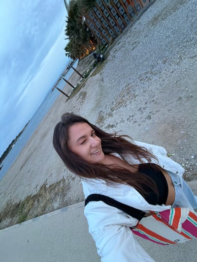 A young woman stands on a gray sidewalk near the beach in Glifada, Greece. She is wearing a black top, light-colored denim jacket, and a white jacket. A multi-colored bag hangs on her shoulder. In the background, there is a body of water, a beach, a palm tree, and a structure with lights. The sky is overcast.