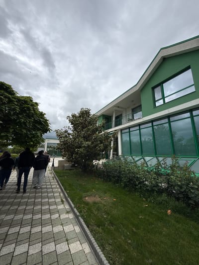 A low-angle shot shows the exterior of a building and a paved walkway, likely located in Gorna Mitropolia, Bulgaria. The building is green and white, with a modern design featuring large glass windows and a balcony with railings. A green roof extends over a covered area. Patches of grass and bushes are visible adjacent to the building. Four people walk along the paved walkway in the foreground. The walkway is constructed of light and dark grey square pavers. The sky is overcast with heavy clouds. Two trees flank the walking path, one on each side.