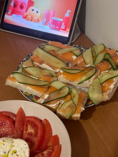 The image shows a close-up of a table with food and a tablet device. On a patterned plate, there are open-faced sandwiches topped with cream cheese, salmon, and cucumber slices. To the left, a white plate holds sliced tomatoes and what appears to be fresh mozzarella cheese. In the background, a tablet displays a cartoon image with pink and orange characters and bottles. The scene is set on a wooden surface, likely a table. This image was captured in Skawina, Poland.