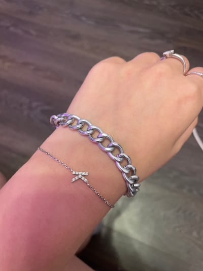 A person's left wrist is shown, adorned with two silver bracelets against a blurred background of dark wood-grain flooring. The upper bracelet is a chunky, curb-link chain. Below it, a finer silver chain bracelet features a small, diamond-encrusted charm shaped like a bow. The skin on the wrist exhibits a warm, reddish hue, possibly from light or a warm undertone. Several rings are visible on the fingers of the same hand: a prominent silver ring with a clear stone on the index finger, and a thinner silver band with small stones on the ring finger. The scene lacks any specific indicators of the city of Romanovka, Russia, or any identifiable urban elements.