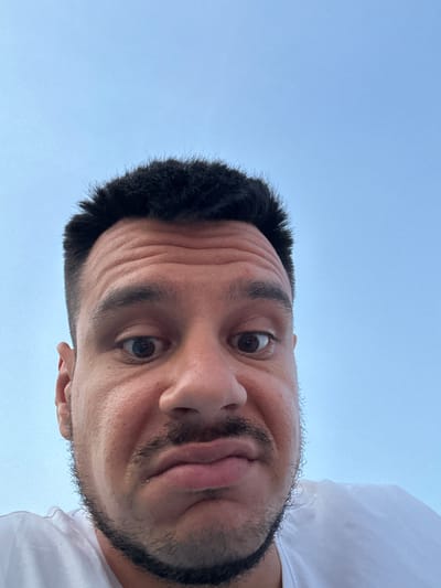 The image is a close-up, low-angle selfie of a person's face against a clear blue sky. The person has dark hair styled upwards, prominent eyebrows, and is making a pouty facial expression with their lips pursed. They appear to be wearing a white t-shirt. The skin shows some texture and what appears to be a light beard growth on the chin and jawline. The background is uniformly blue, suggesting an outdoor setting during the day. There is no discernible information within the image that would indicate a specific location in El Parador de las Hortichuelas, Spain.