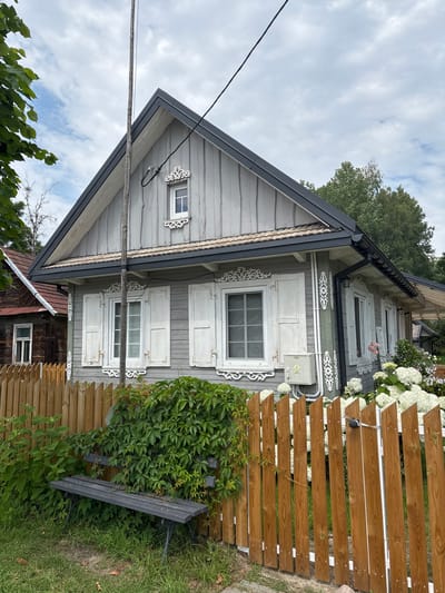 The image depicts a traditional wooden house in Trześcianka, Poland. The house is painted gray with white shutters and decorative trim around the windows. A wooden fence encloses the property, with a lush green vine growing over a dark-colored bench placed near the fence. In the background, a neighboring wooden house with a red roof is visible, along with trees and a cloudy sky. The scene appears to be a quiet, rural setting.