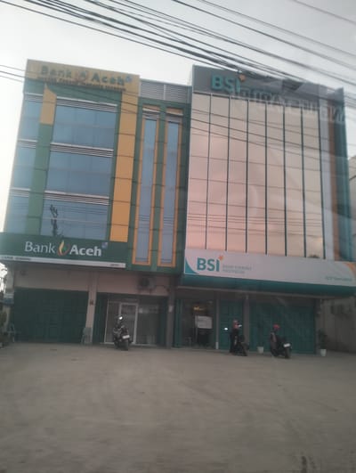 This media file shows the exterior of two adjacent bank buildings in Blangpeuria, Indonesia. The building on the left is the "Bank Aceh" and the building on the right is "BSI BANK SYARIAH INDONESIA". Both buildings have multiple stories with large windows. The Bank Aceh building has a green and yellow facade, while the BSI building has a modern glass facade. In front of the buildings, there are several parked motorcycles. The ground in front of the buildings appears to be a paved area. The sky is visible at the top of the image, with some electrical wires crossing the frame. The overall lighting suggests it is daytime. The scene depicts a typical street view of financial institutions in a commercial area. Visible text includes the names of the banks, "Bank Aceh", "BSI", "BANK SYARIAH INDONESIA", and the branch information "KANTOR CABANG PEMBANTU GEUDONG", "CAPEM GEUDONG", and "KCP Samudera". There is also "atm" visible below the Bank Aceh signage.