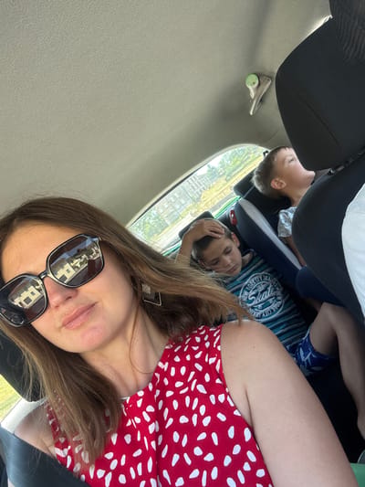 This media file is a vertical, low-angle selfie taken from inside a vehicle, likely a car. The subjects are two young boys and a woman. The boys are seated behind the woman, who is in the foreground. The boy in the middle is resting his head on his arm, and the boy behind him is looking towards the side. The woman is wearing a red and white polka dot dress and sunglasses. Her hair is brown and falls over her shoulders. In the reflection of her sunglasses, buildings are visible, suggesting an urban environment.
The scene appears to be a road trip, with the children likely being passengers. The lighting suggests it is daytime. The overall mood seems casual and relaxed, typical of a family outing.
The setting is the interior of a vehicle, with glimpses of the outside through the window. The window shows green foliage and some buildings. The interior of the car includes a black headrest and a grey interior panel. A green object, possibly a drink or a small toy, is attached to a silver handle on the grey panel.
There is visible text on the blue and white striped t-shirt worn by the boy in the middle. It reads "NEW YORK CITY SKATE". The woman's sunglasses have a reflective surface, showing a blurred image of buildings.