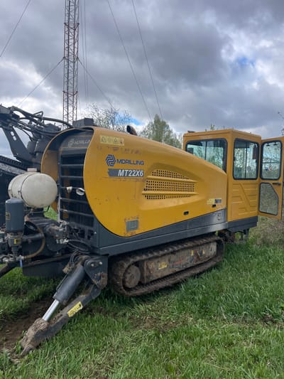 A yellow horizontal directional drilling rig, labeled "MDRILING MT22X6," sits in a grassy field. The rig has a tracked chassis and an open cab. A crane structure is visible in the background under a cloudy sky. The ground is a mix of grass and dirt, showing signs of recent activity.