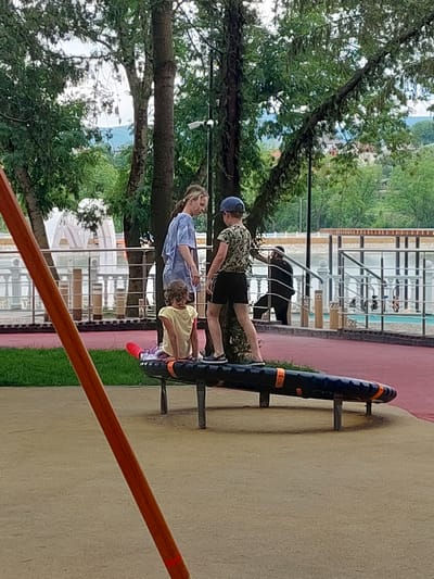 The image shows a playground area in Alikonovka, Russia, with three people near a seesaw-like play structure. A woman in a blue shirt stands to the left, looking down at a young child wearing a yellow shirt and patterned leggings who is sitting on the play structure. To the right of the woman, a child wearing a camouflage t-shirt and dark shorts, with a blue baseball cap, is standing on the same play structure, facing away from the camera. In the background, there is a fence with metal railings, a body of water, and trees. The ground in the foreground is a mix of light brown dirt and red surfacing.