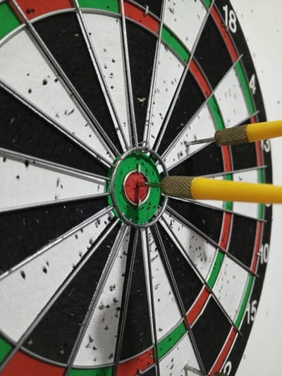 The image presents a close-up, slightly angled view of a standard bristled dartboard. The board features a traditional layout of twenty numbered segments, alternating black and white, defined by silver-colored wire dividers. Concurrently, red and green rings denote double and triple scoring zones, with a central red bullseye and an outer green double-bull. The surface of the dartboard shows numerous small punctures, consistent with repeated impact from darts. Two yellow-shafted darts are embedded in the board. One dart is precisely lodged in the central red bullseye. A second dart is positioned in the green double-bull ring, slightly above and to the right of the bullseye dart. Both darts possess metallic, knurled grips. The dartboard is mounted against a plain, light-colored background, suggesting an indoor environment. This scene is located in the city of Al Haouafate, Morocco.