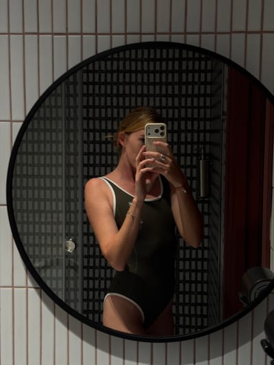 A woman is taking a mirror selfie in a bathroom. She is wearing a dark green one-piece swimsuit with white trim. She is holding a smartphone in her right hand, which is raised to capture the reflection. She has light brown hair pulled back and is wearing a ring on her left hand and a bracelet on her right wrist. The bathroom has white tiled walls with thin pink grout lines. The mirror is round and has a black frame. In the reflection of the mirror, a black and white patterned tile wall is visible behind the woman, along with a chrome soap dispenser and a shower door. The overall impression is that of a modern, clean bathroom. There is no indication that this location is in Kaji-Say, Kyrgyzstan.