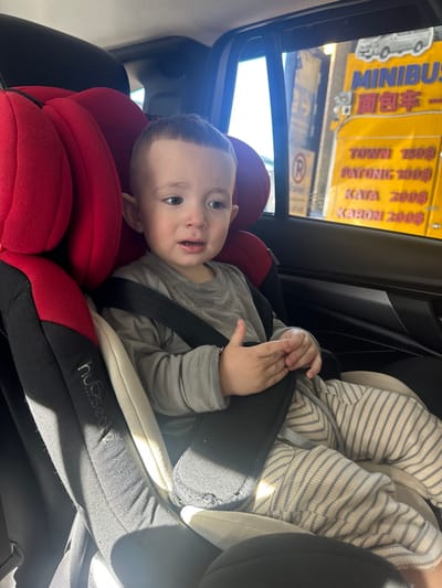 A young child, secured in a red and black car seat, faces the camera with a slightly open mouth. The child wears a gray long-sleeved shirt and striped pants, and is strapped in. Outside the car window, reflections of a yellow sign with writing can be seen. The car interior features black panels and a visible sunroof.