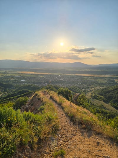 The image displays a panoramic landscape captured from a high vantage point near the city of Stob, Bulgaria, at sunset. In the immediate foreground, a well-defined dirt trail, composed of reddish-brown soil and small rocks, curves gently from the lower left towards the mid-right. The path is flanked by patches of dry, golden-brown grasses and various green shrubs, some exhibiting small yellow blossoms. The terrain on the steep slopes adjacent to the path appears eroded and rocky. In the midground, the valley floor below reveals the clustered buildings of the city of Stob, interspersed with green trees and cultivated fields. Beyond the city, a mosaic of agricultural fields, ranging from dark green to lighter golden-brown, stretches across the valley, enclosed by a distant horizon line. Multiple layers of hazy, undulating mountain ranges are visible in the background. The sky above is characterized by a setting sun positioned in the upper-center, emitting a bright golden light and casting a warm glow across scattered clouds that show both illuminated edges and darker undersides. The upper portion of the sky is a clear, pale blue, transitioning to warmer tones near the horizon. No individuals or specific actions are discernible within the scene.