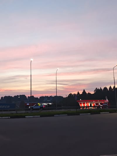 This is a low-angle, eye-level, medium shot of a gas station with a red and white awning at dawn or dusk. The sky is a soft pink and blue gradient, with thin, wispy clouds. Several streetlights are visible, casting a warm glow. In the middle ground, a white SUV is parked at the gas station. There are also buildings and trees in the background. The foreground is a dark asphalt road with a black and white striped curb. The scene appears to be set in Dubrovki, Russia.