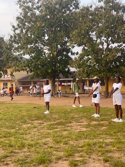 The image captures a group of young adults, seemingly participating in some form of outdoor activity or training, in Emure-Ekiti, Nigeria. The setting appears to be an open field with dry, sparse grass and patches of dirt. In the background, there are several low-rise buildings with yellow and orange facades, suggesting a school or institutional campus. Some buildings have visible laundry hanging out to dry.

A volleyball net is set up across the field, and a few individuals are engaged near it, possibly playing or preparing to play. Several individuals in the foreground and middle ground are dressed in white athletic wear, consisting of shorts and t-shirts or polos, with some wearing white caps. They appear to be standing in formation or following instructions. One woman in the immediate foreground, facing the camera, is also in white athletic attire and seems to be observing or participating in an activity. Another individual in the middle ground, wearing a green military-style uniform and white shorts, is also present. The overall atmosphere suggests a structured activity, possibly a physical training session or a sports event. The presence of a large tree with green foliage in the foreground frames the scene. The sky is overcast, and the lighting suggests it is either early morning or late afternoon.