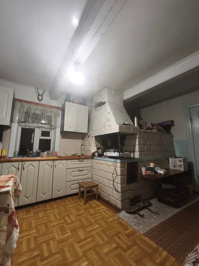 The image depicts a dimly lit, rustic kitchen in what appears to be a rural home, possibly in the Навадворскі сельскі Савет region of Belarus. The room features a large, tiled oven or fireplace dominating one side, with a light-colored tiled facade and a white, stuccoed hood. Above the oven, shelves are cluttered with various items, including bottles, containers, and what looks like fabric or bags.
The kitchen cabinets are a pale, off-white color with decorative molding, and they run along one wall, meeting a sink area beneath a window. The window itself is covered by a sheer, patterned curtain and reveals a dark exterior. Above the window, a mounted deer head with antlers serves as a decorative element. The countertop area has several items on it, including jars, mugs, and what appears to be a bottle of cleaning product.
The floor is covered in a parquet-style linoleum in shades of brown and yellow. A small wooden stool is positioned near the cabinets. On the right side of the frame, a wooden table holds a blender box and other assorted objects. The ceiling is wooden and has exposed beams, with a fluorescent light fixture providing illumination. The overall impression is one of a lived-in, traditional dwelling. There are no people visible in the image.