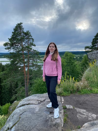 A young woman with long, brown hair stands on a rocky outcrop overlooking a serene landscape under a cloudy sky. She is wearing a pink hooded sweatshirt with a white turtleneck peeking out, black pants, and white sneakers. Her gaze is directed forward, toward the camera. The background features lush green trees, a body of water, and a distant, rolling landscape. The rocky terrain beneath her feet is rough and uneven, with patches of grass and wildflowers growing among the stones. The overall atmosphere is one of natural beauty and tranquility. The location is Khelyulya, Russia, a place known for its picturesque natural surroundings and recreational opportunities.