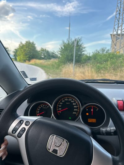 The image shows a view from the driver's seat of a car, looking towards the dashboard and the road ahead. The car appears to be on a paved road with a gentle curve, bordered by dry grass and trees. In the background, a utility tower and a building are visible against a bright, cloudy sky. The dashboard displays the car's speed and fuel gauge, with the odometer showing "30% 2275". The steering wheel is visible in the foreground, with the Honda logo at its center. The context of Novo Zhelezare, Bulgaria is provided but not visually evident in the image.
