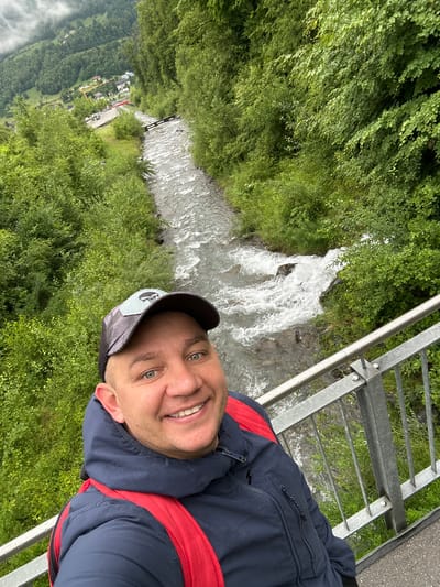 A selfie is taken by a man standing on a bridge with a metal railing. He is wearing a dark blue jacket with a red backpack underneath and a baseball cap. The man is smiling at the camera. Behind him, a river flows rapidly through a lush, green, mountainous landscape. Trees and vegetation line the riverbanks. In the distance, a small town or village is visible nestled in the valley, with buildings and some construction equipment. The sky is overcast, suggesting a cloudy day. The location is the city of Hüsen, Switzerland.