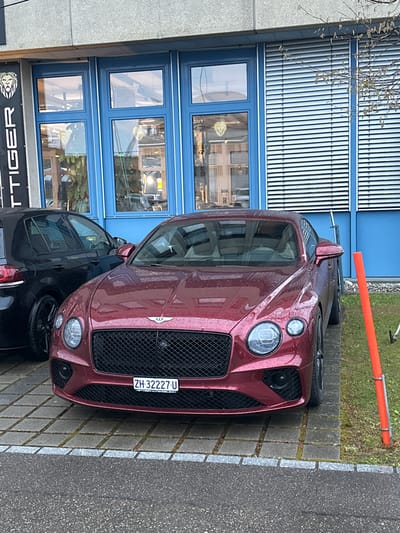A front-facing view depicts a maroon Bentley car parked on a paved surface adjacent to a building with blue-framed windows and a gray exterior wall in Wetzikon (ZH), Switzerland. The license plate is visible on the car, reading "ZH 32227-U". A black car is partially visible parked behind the Bentley. A sign displaying the word "TIGER" is also visible. There is grass and a orange pole behind the Bentley.