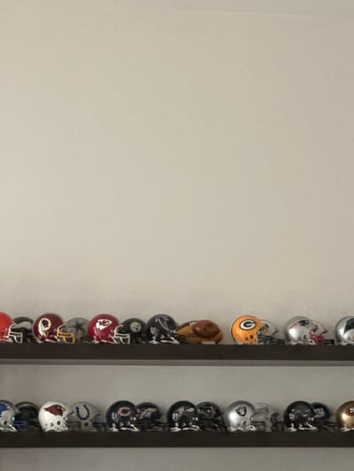 The image depicts an indoor scene in McAllen, United States of America, showcasing two horizontal shelves against a plain, light-colored wall. Both shelves are dark-toned and display a collection of miniature National Football League (NFL) helmets.
On the top shelf, from left to right, the visible objects include: a partially visible red helmet; a black helmet with an indistinct silver logo; a burgundy helmet with a yellow 'R' and feather emblem; a silver helmet with a blue star; a red helmet with a white arrowhead logo; a black helmet with a yellow 'Steelers' logo; a black helmet with a silver falcon emblem; a brown, football-shaped object; an orange helmet with a green 'G' logo; a silver helmet with a red and blue patriotic logo; and a partially visible silver helmet.
On the bottom shelf, from left to right, the visible objects include: a blue helmet with a white stylized 'NY' logo; a white helmet with a red cardinal bird logo; a blue helmet with a white horseshoe logo; a dark blue helmet with an orange 'C' logo; a dark blue helmet with a red 'NY' logo; a black helmet with a silver 'R' logo; a silver helmet with a white ram's horn logo; a black helmet with an indistinct white stylized logo; a blue helmet with a white 'Bills' logo; and a partially visible gold helmet.
No individuals, actions, or interactions are present in the static display. The environment consists solely of the wall and the two shelves holding the miniature helmets.