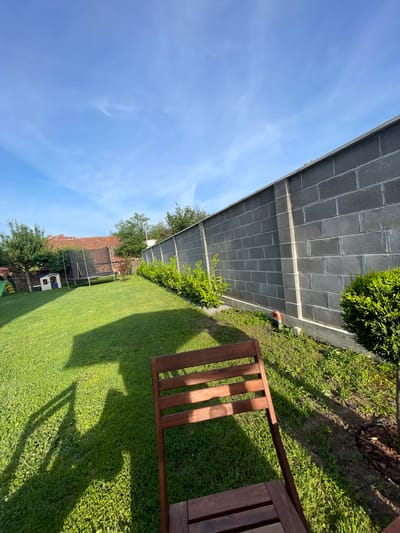 The image depicts a backyard in the city of Levski, Bulgaria. A grey brick wall borders the yard on one side, with greenery and shrubs growing along its base. The yard consists of a green lawn. A wooden chair is partially visible in the foreground. In the background, a trampoline and a small playhouse can be seen, along with other trees and structures. The sky is blue with some clouds.