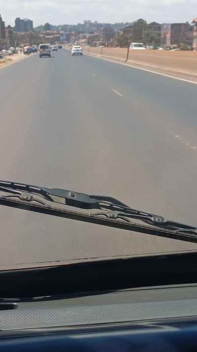 The image captures a forward-facing view from inside a vehicle on a multi-lane highway in Kinoo ward, Kenya, under overcast daylight conditions. In the immediate foreground, a dark grey dashboard and a black windshield wiper assembly are visible. The central and mid-ground depict a wide, grey asphalt highway with faint white dashed lane markings. Multiple vehicles are present in the distance, including a light-colored SUV in the left lane and a white sedan further ahead in the right lane, both traveling away from the camera. To the left of the main highway, a shoulder or service road is visible with additional vehicles and multi-story buildings interspersed with sparse vegetation, indicative of urban development. To the right, a continuous concrete barrier separates the highway from an adjacent area where a white van is partially visible behind the barrier, with more buildings in the background. No specific individuals are discernible in the scene.
