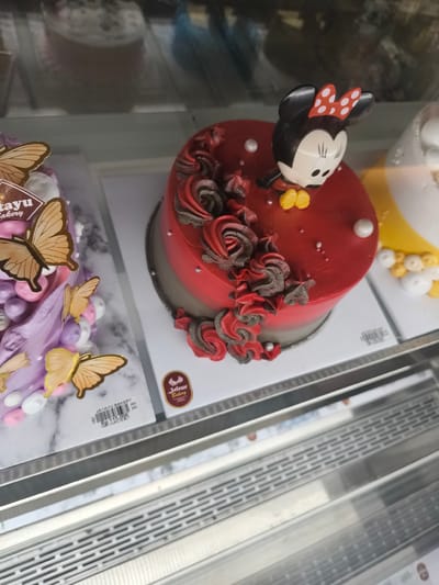 Two cakes are displayed in a refrigerated bakery case. The cake on the left is decorated with purple frosting, edible butterflies, and light purple and pink candy. The cake on the right is red and decorated with dark red and gray frosting swirls and a Minnie Mouse figurine on top. A label for "Jatayu Bakery" is visible on the cake on the right. Another label for "Jatayu Bakery" and a price of "Rp 115.000" are visible on the cake on the left. The bakery case is made of metal and glass. The background is blurred and shows reflections of other items in the bakery.