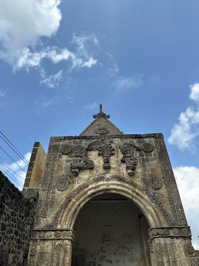 The image shows a low-angle, medium shot of the facade of a stone structure in San Andrés Calpan, Mexico. The facade is ornate, with intricate carvings. Above an arched entryway, there are relief carvings of what appear to be figures and decorative elements, including what looks like a tree or plant. The stone is weathered and aged. To the left of the facade, a wall constructed of dark, rough stones is visible. Above the structure, a blue sky with scattered white clouds is present. Power lines stretch across the upper left portion of the image. The overall impression is one of historical architecture against a natural sky.