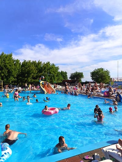 This is a full-shot, eye-level, outdoor photograph taken on a sunny day at the "Bochkari" water park in Yarovoye, Russia. The image captures a vibrant scene of a crowded swimming pool filled with people of various ages enjoying themselves. The water is a clear, bright blue, and the pool is surrounded by a blue deck.
In the foreground, several individuals are visible. On the left, a man is partially submerged in the water, facing away from the camera. To his right, a young boy with short brown hair is excitedly looking towards the camera, his mouth open. Further to the right, a young girl with dark hair and glasses is playing in the pool, wearing a pink swimsuit.
In the middle ground, more people are scattered throughout the pool. A woman in a pink bikini is wading, and a child in a yellow swimsuit is swimming nearby. A girl is seen floating on a pink donut-shaped inner tube, looking to her left. Children are also playing on a water slide structure designed to resemble a pirate ship, with a red slide and various colorful figures.
In the background, numerous people are lounging on white beach chairs, many of them under umbrellas. Trees line the perimeter of the water park, and a building with signage that includes "SAUNA" and a logo with lemons is visible on the left. The sky is a bright blue with scattered white clouds.
The overall atmosphere is one of lively recreation and summer enjoyment.