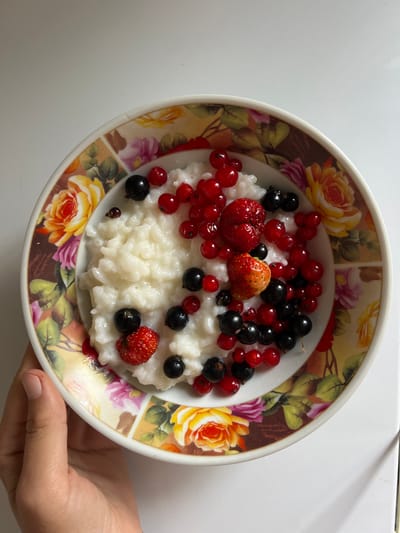 A close-up, top-down shot shows a bowl of rice pudding adorned with fresh red currants, black currants, and two small strawberries. The bowl itself features a floral pattern with roses in shades of yellow, pink, and orange against a brown background. A human hand is visible holding the bowl from the left. The scene is set against a plain white background, likely a tabletop. The location is not specified in the image, therefore it cannot be determined to be Pivkino, Russia.