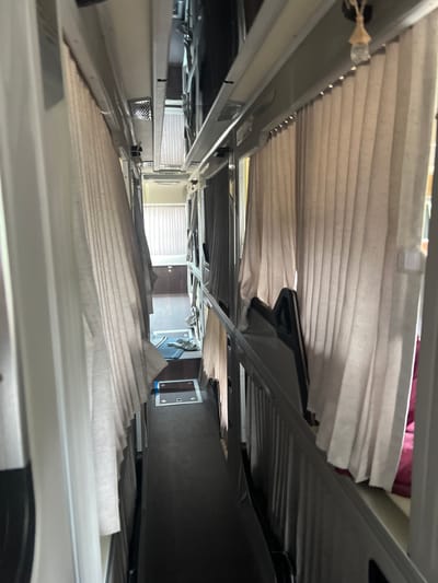 The image depicts an interior space, possibly within a vehicle or a confined structure in Đăk La, Vietnam. The perspective is from a low angle, looking upwards towards a series of overlapping, pleated blinds or shades. The blinds are primarily a light beige or off-white color. The structure supporting the blinds appears to be metallic, with a silver or gray finish. The lighting suggests an interior setting. In the background, a window with partially drawn horizontal blinds and a doorway are visible. The room beyond is indistinct but seems to contain dark furniture and wall decoration. The blinds appear to be folded and close together, with spaces between them.